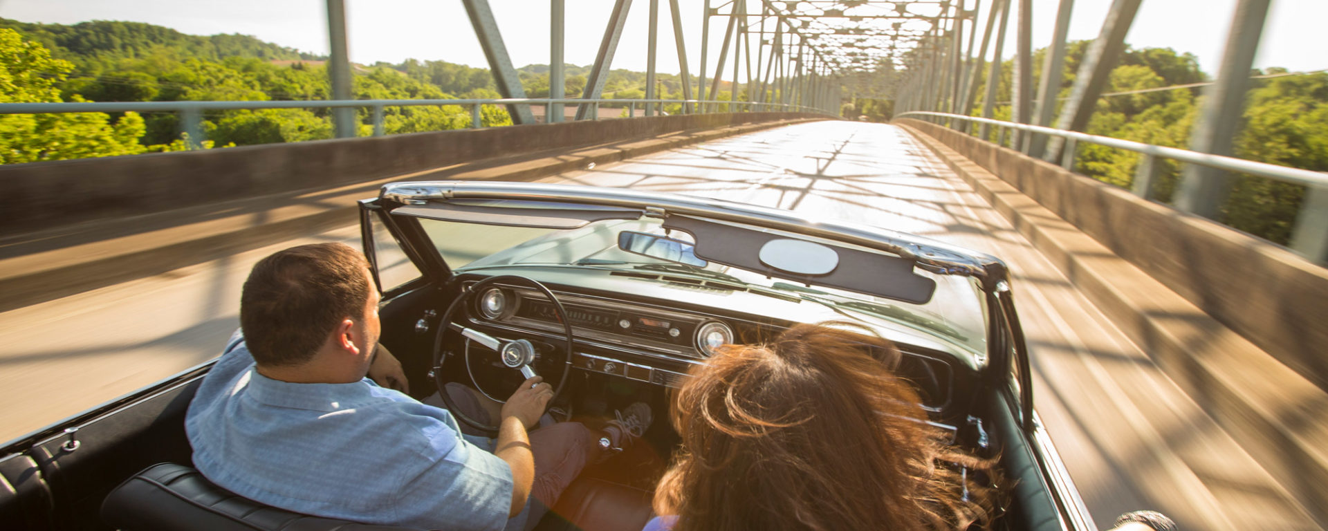 A man and woman cross a river bridge in an open-top convertible on a sunny day