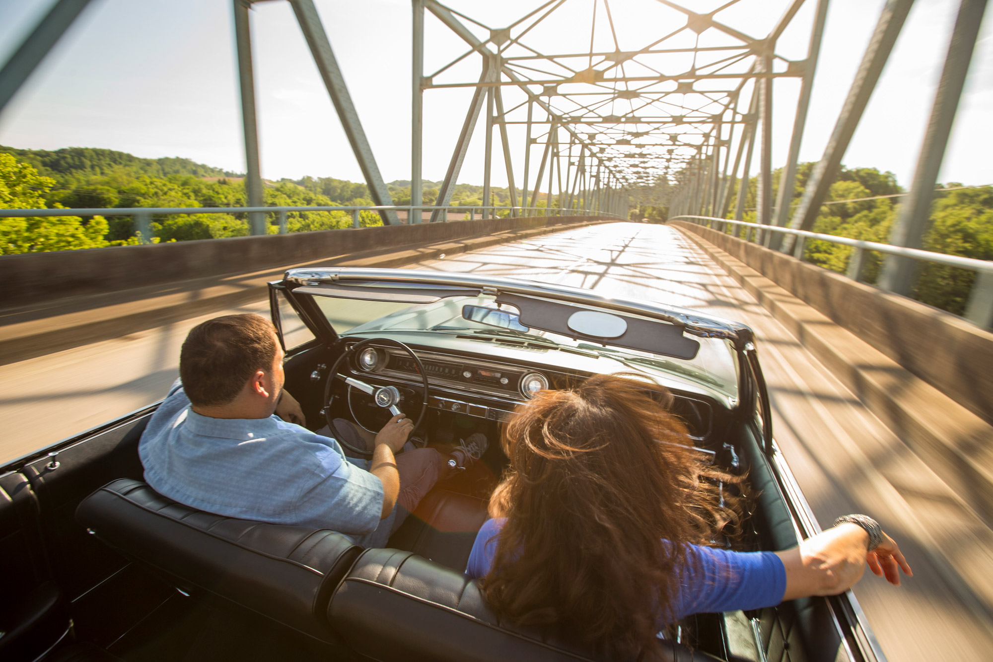 A man and woman cross a river bridge in an open-top convertible on a sunny day