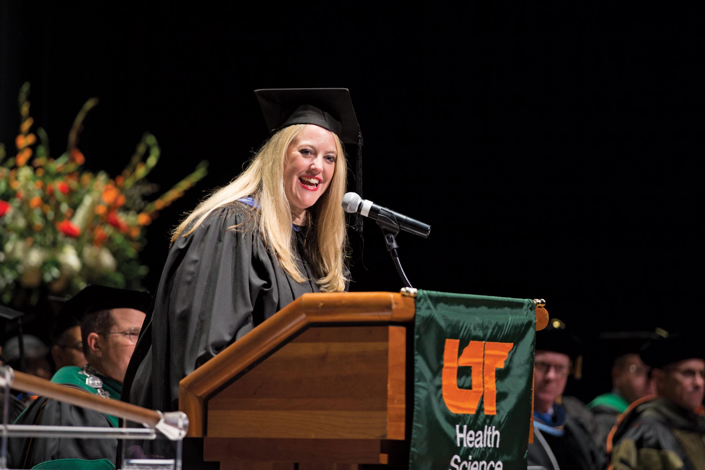 Betsy Brasher Melby wearing black robe and mortarboard cap speaks at commencement podium