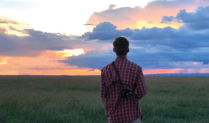 Austin Holdsworth stands outdoors facing a sunset with a camera strap hanging off his right shoulder