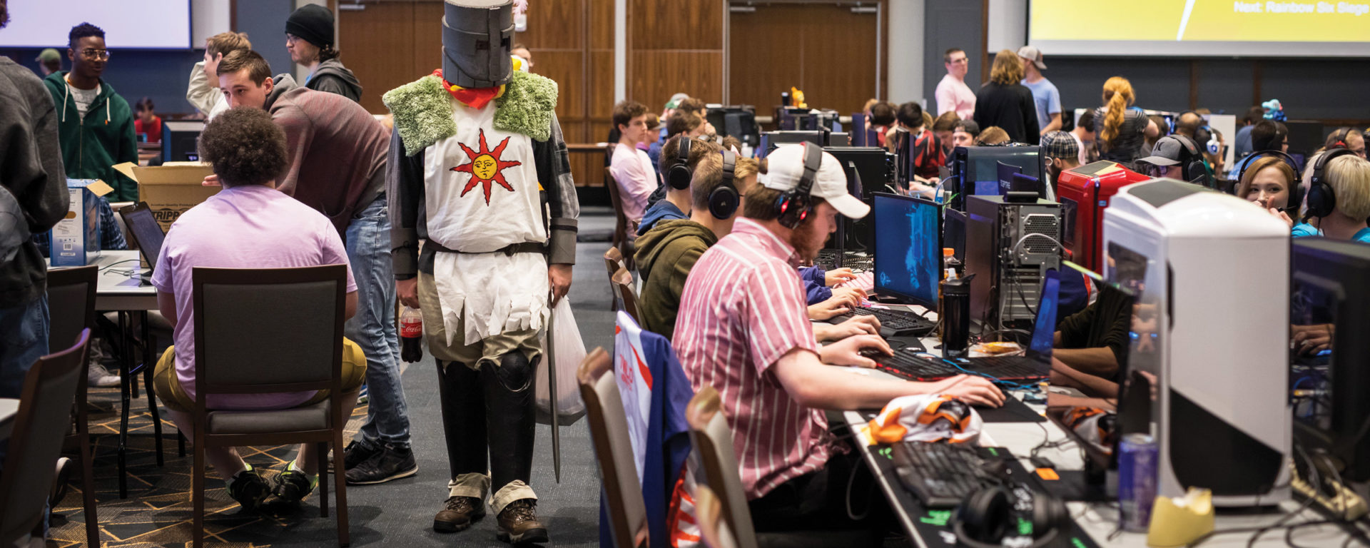 A student dressed as a knight walks between rows of gamers at PC consoles