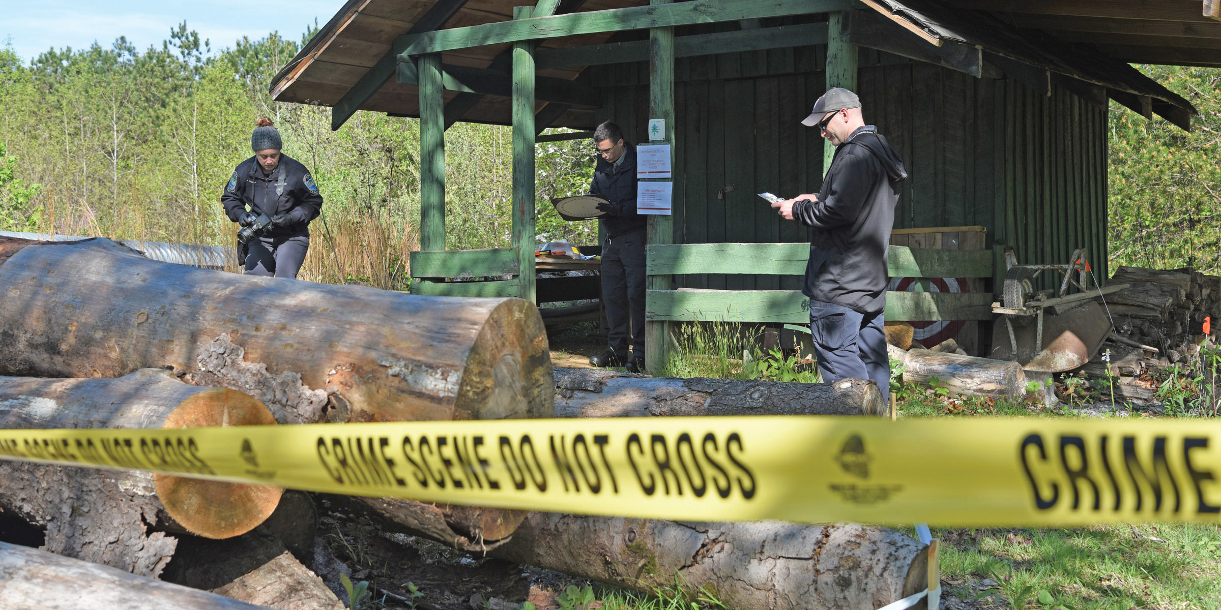 technicians in tactical gear photograph a shed behind yellow crime scene tape