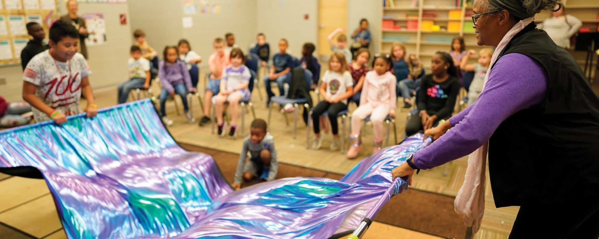 A student and teacher wave a rainbow tie-dyed banner in elementary school music class