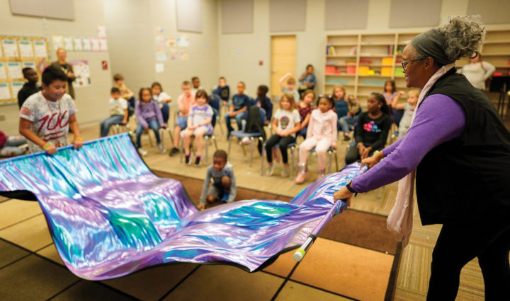 A student and teacher wave a rainbow tie-dyed banner in elementary school music class