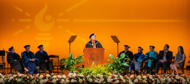 Donde Plowman speaks at a podium flanked by UT officials in academic regalia