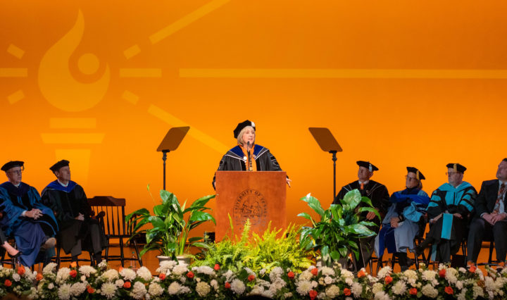 Donde Plowman speaks at a podium flanked by UT officials in academic regalia