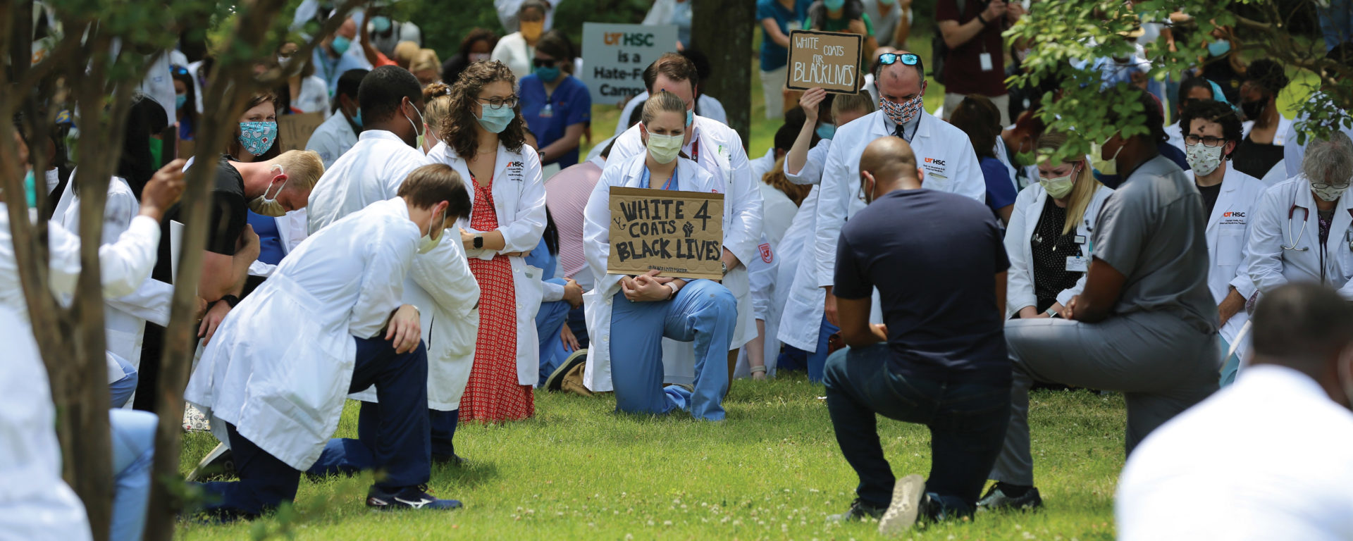 People of all races in white coats and scrubs kneel in the grass, wearing face masks and holding protest signs
