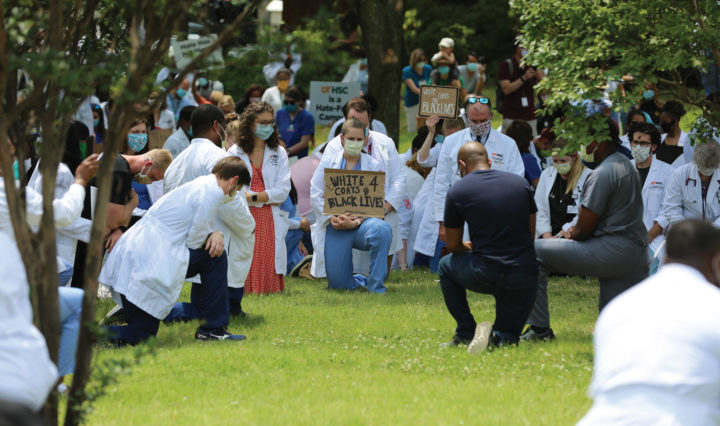 People of all races in white coats and scrubs kneel in the grass, wearing face masks and holding protest signs