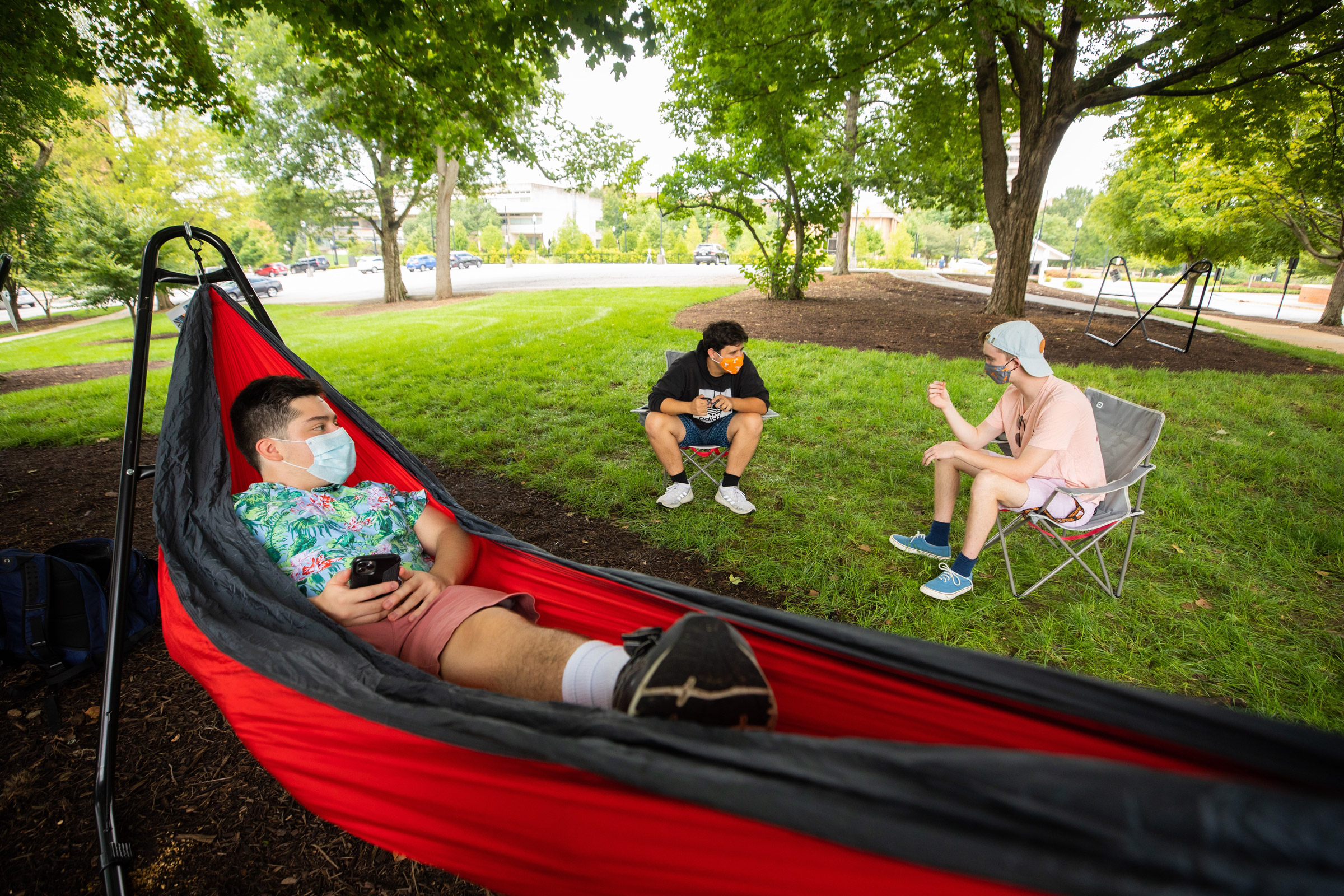 Three men chat outdoors in the shade wearing face masks. One is in a hammock and two are seated in lawn chairs.