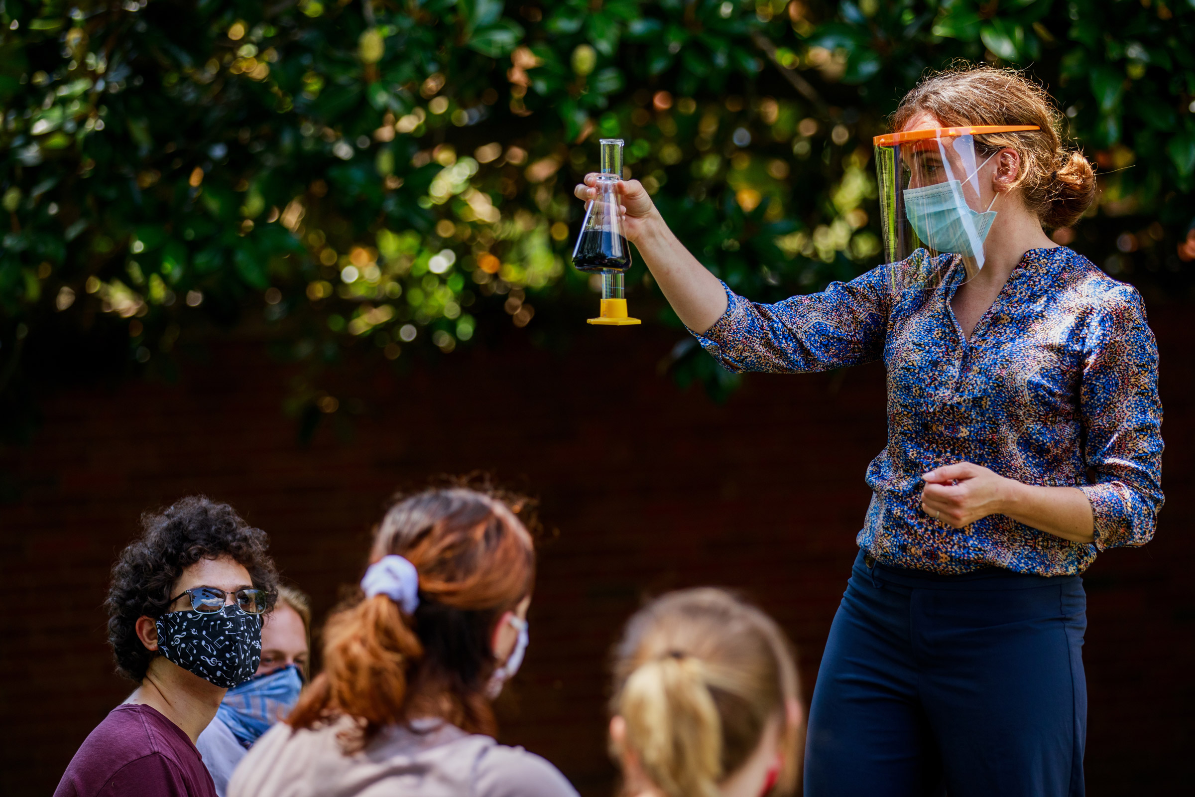 A chemistry professor in a mask and face shield raises a beaker filled with a liquid during an oudoor demonstration