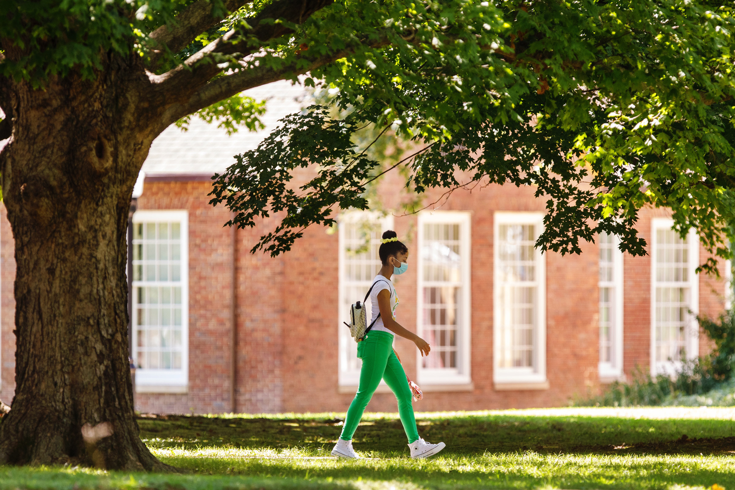 A woman in bright green pants and high top sneakers walks under a shaded path to her class