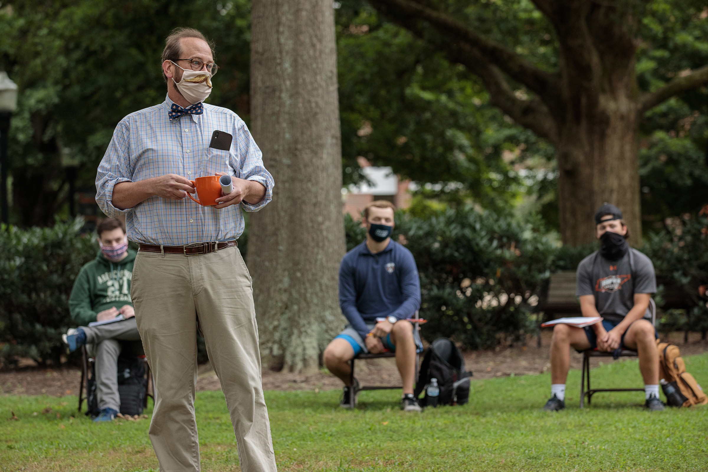 A philosophy professor lectures outdoors, wearing a bowtie and a face mask with a false mustache