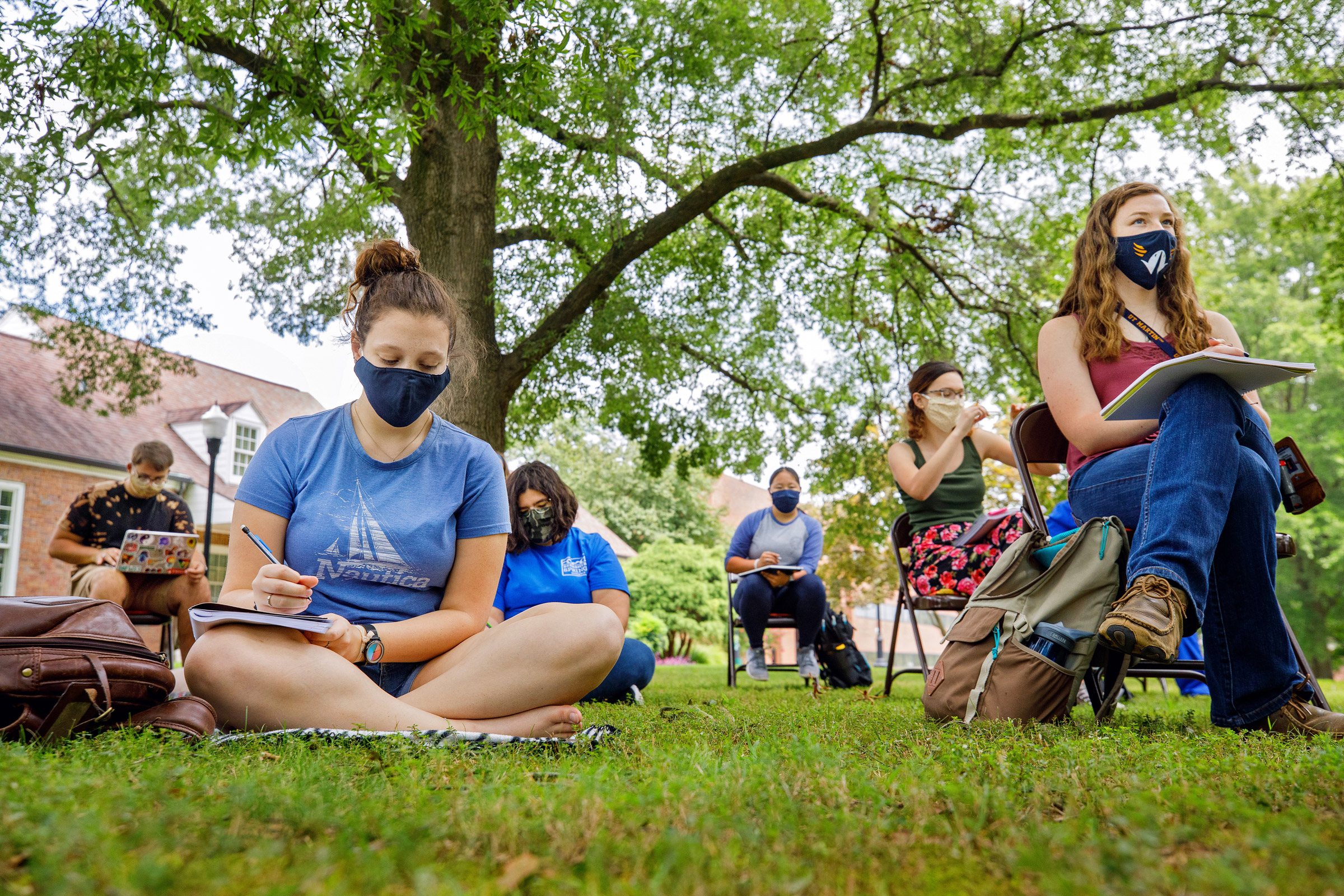 A group of students take notes in the shade outdoors, spaced apart by 6 feet