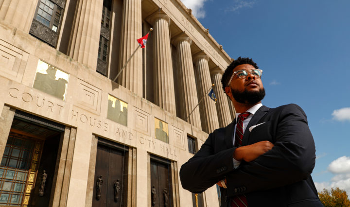 A Black man in a suit and glasses stands in front of the Nashville City Hall with his arms crossed