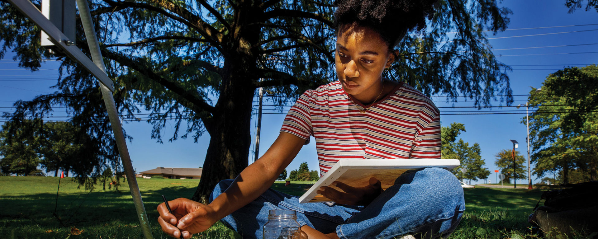 A Black woman sits under a tree and paints on a canvas in an outdoor art class