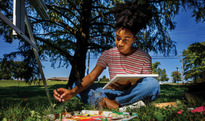A Black woman sits under a tree and paints on a canvas in an outdoor art class