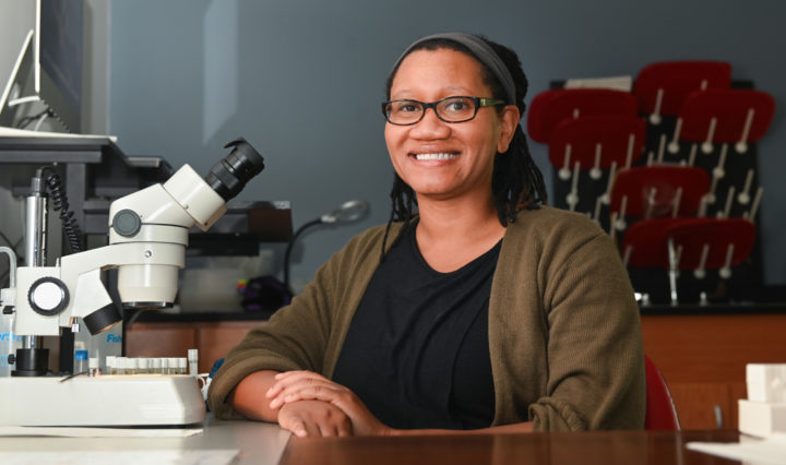 A Black woman seated in front of a microscope smiles confidently in her research lab