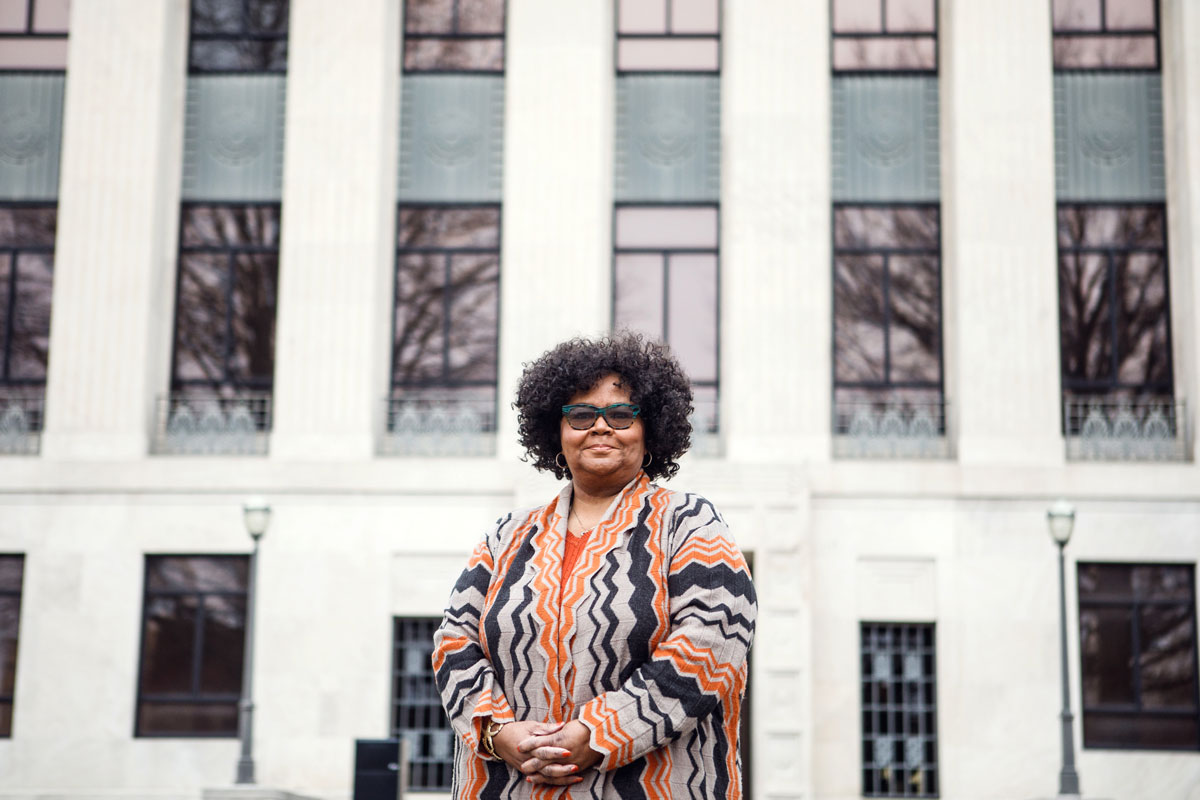 A Black woman stands outside a white civic building in Weakley County. She is dressed in orange and navy and wears progressive lenses in her turqoise glasses