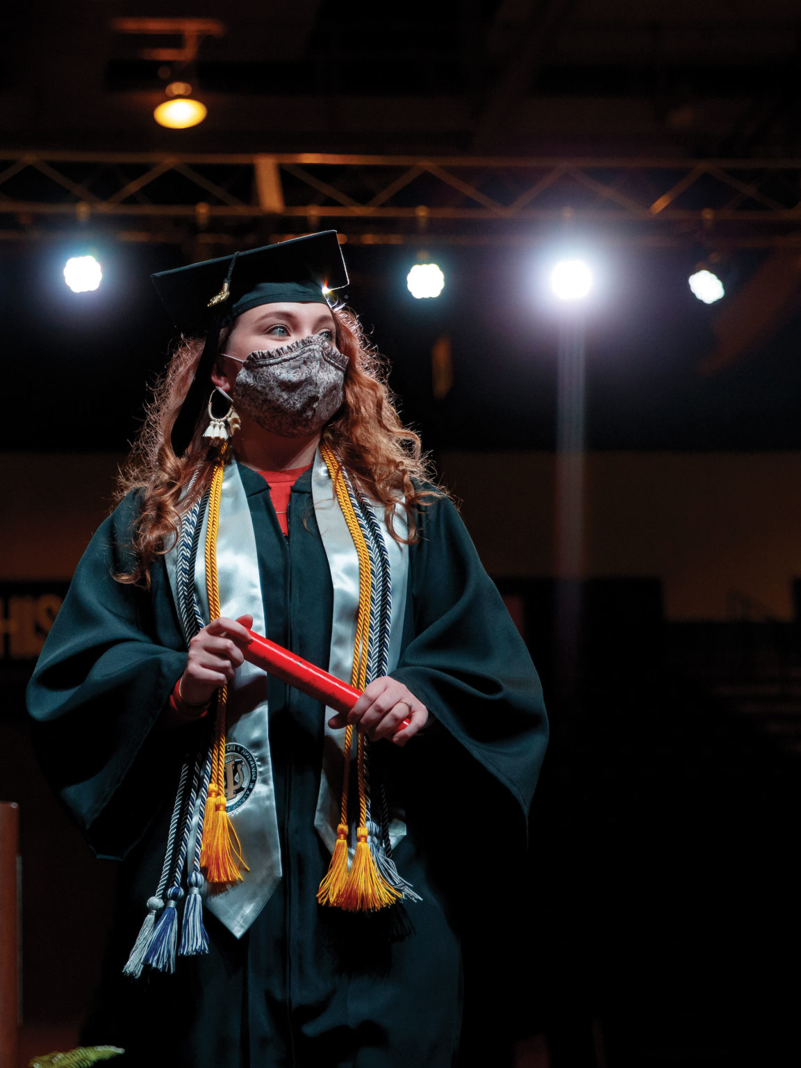 A woman in a cap, gown and face mask walks across a graduation stage with her diploma in hand