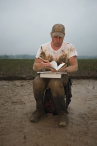 A man sits on a 5 gallon bucket, taking notes in a rice paddy. He is covered in mud from torso to his boots