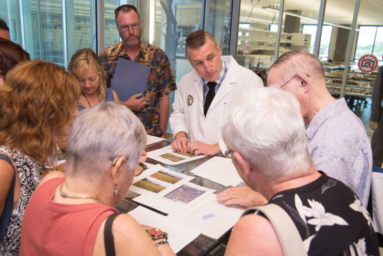 A group of visitors look at photos on a table as Kyle McCormick, dressed in a white lab coat and tie gives a lecture