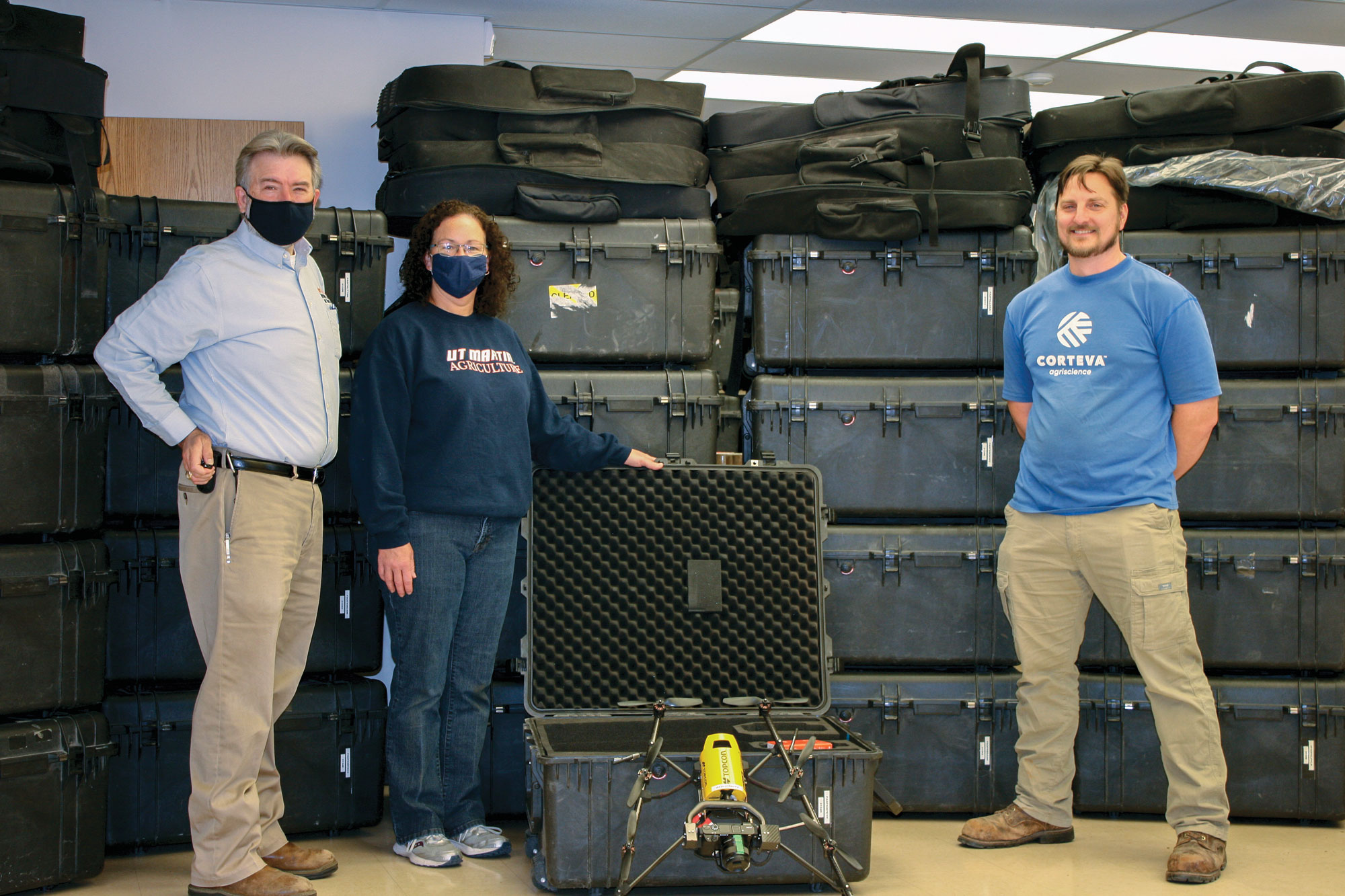 Three people stand in front of stacks of cases of aerial drone equipment
