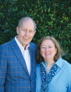 A smiling couple wearing blue, picturedd in front of a holly bush