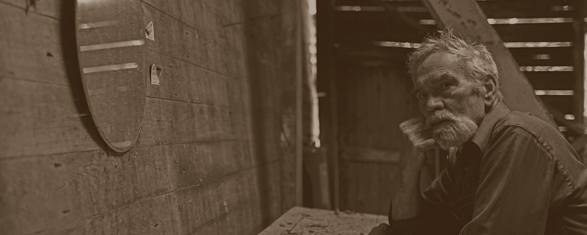 An older man leans on his elbow against a tabe in a barn