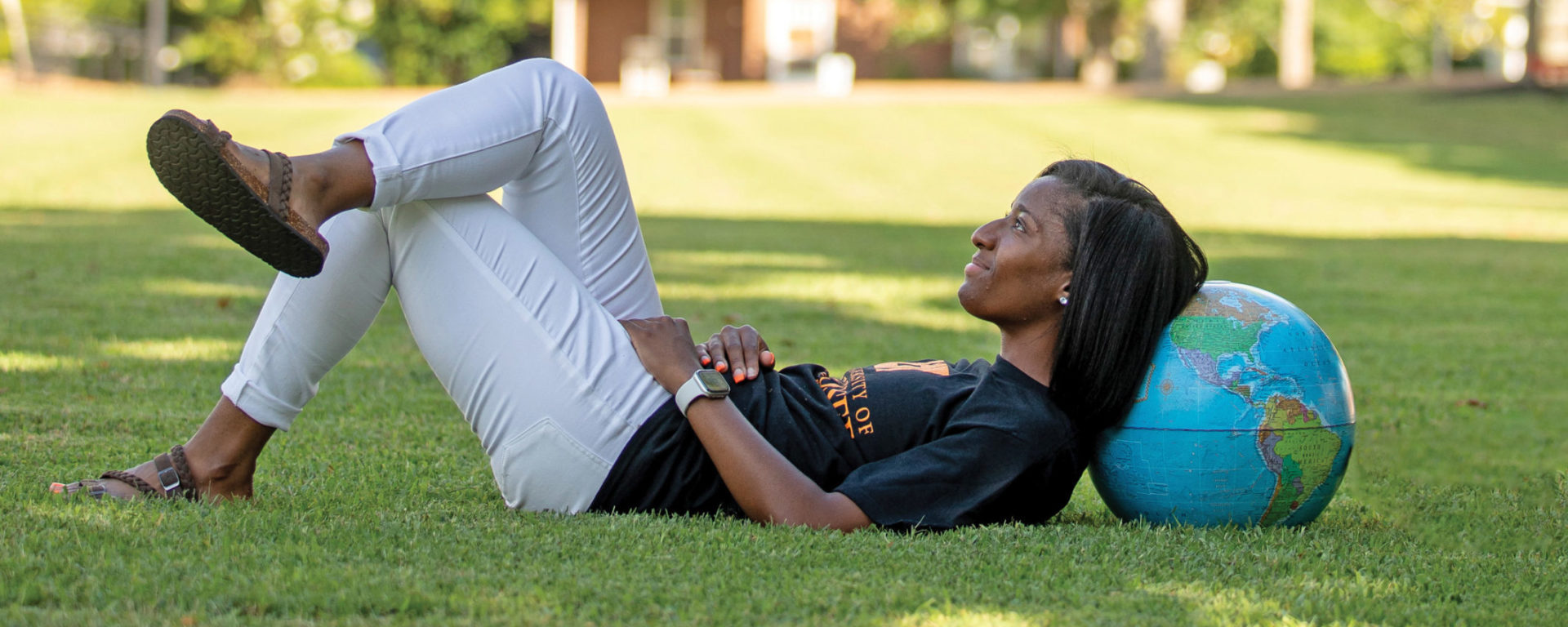 A young black woman rests her head on a globe on a college campus lawn. She is dressed in a black tee shirt, white pants and leather sandals