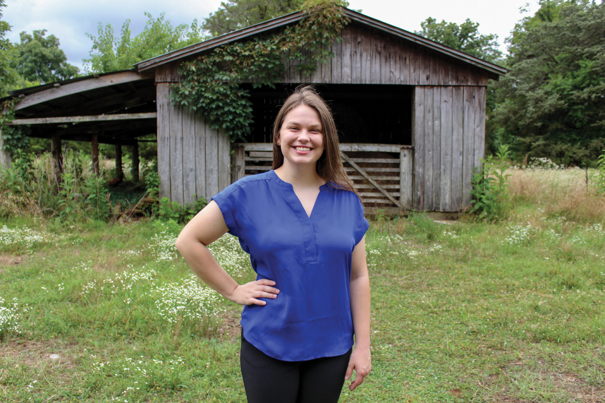 A young white woman in a blue silk shirt stands in front of an ivy covered barn