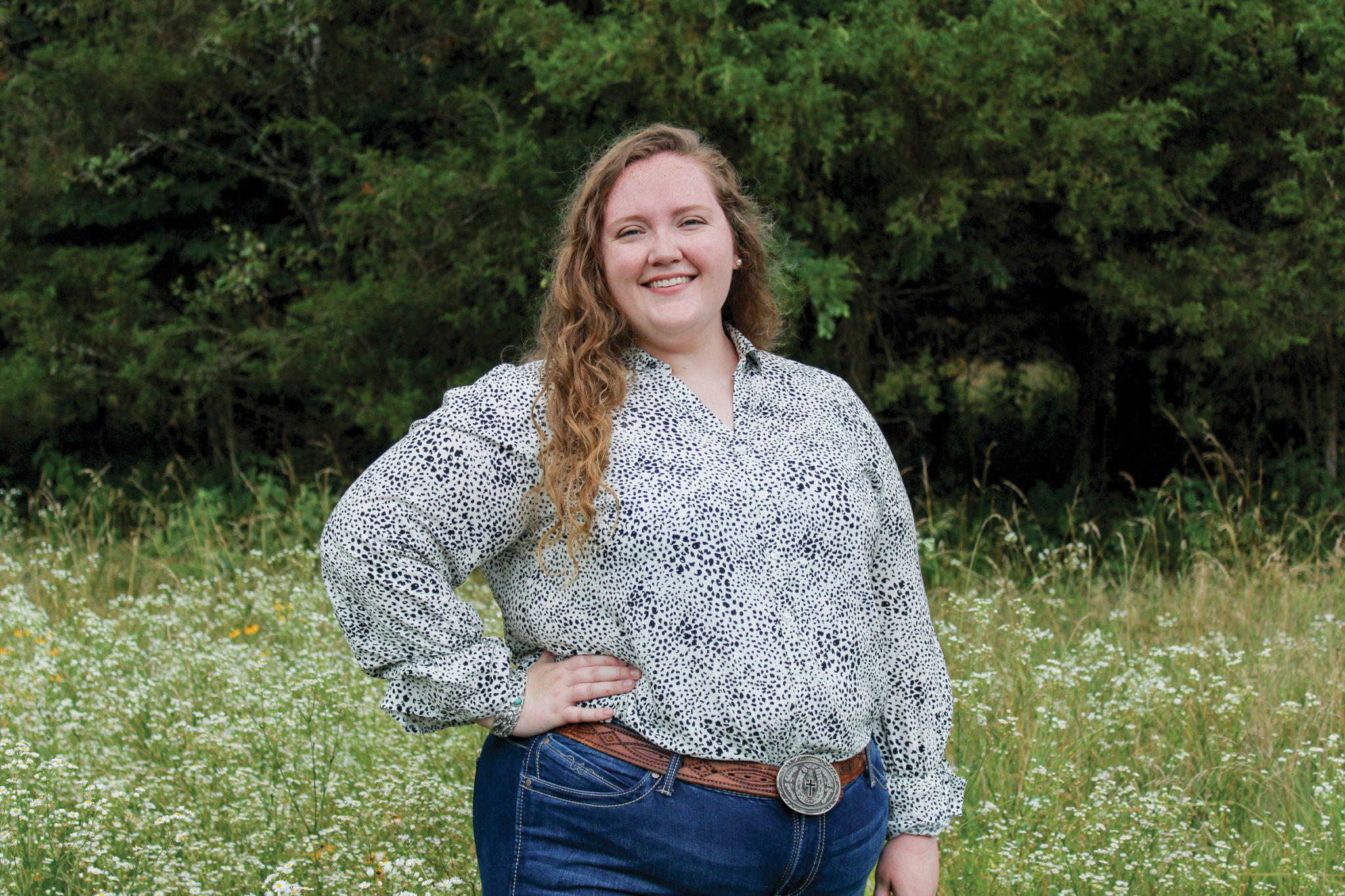A young woman dressed in denim jeans and a floral shirt stands in front of evergreen trees in an overgrown meadow