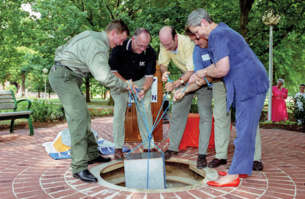 five people lower a metal box into a hole in brick-paved ground
