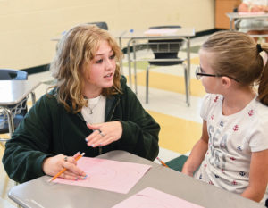 an older teenage girl works with a younger grade school age girl in a classroom setting