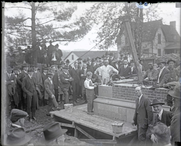 An antique photo shows a young boy setting in a cornerstone carved 1919. A group of men, most wearing hats watch.