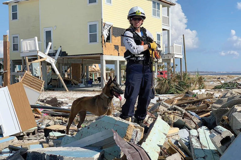 A woman wears a helmet and protective gear surrounded by debris near a collapsed house in the aftermath of a hurricane