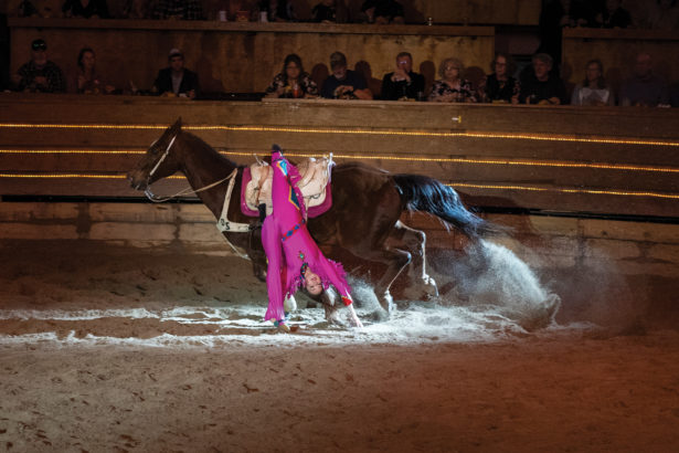 A woman in a bright pink suit hangs unpside down on the side of her horse, performing in an arena