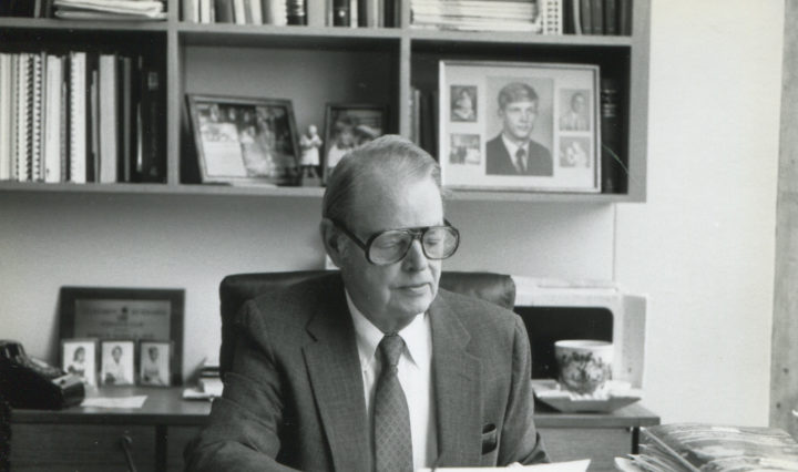 A middle aged man in thick glasses and a tie sits in an office decorated with bookshelves and photos