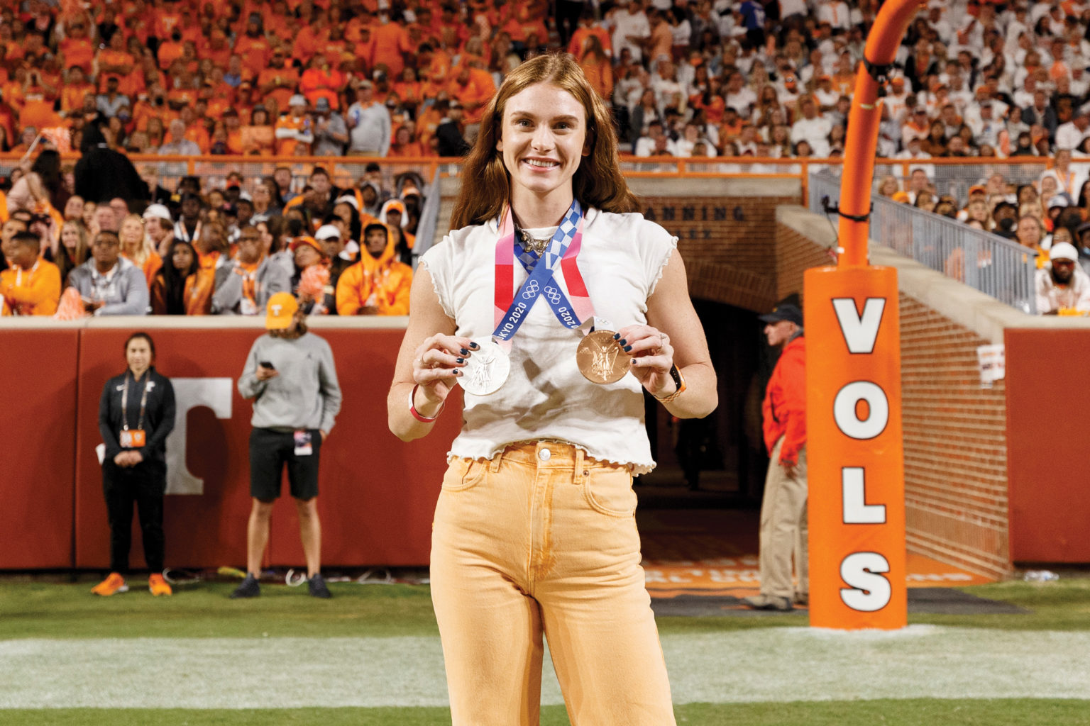 A woman in orange jeans displays her medals worn around her neck on the football field at Neyland Stadium