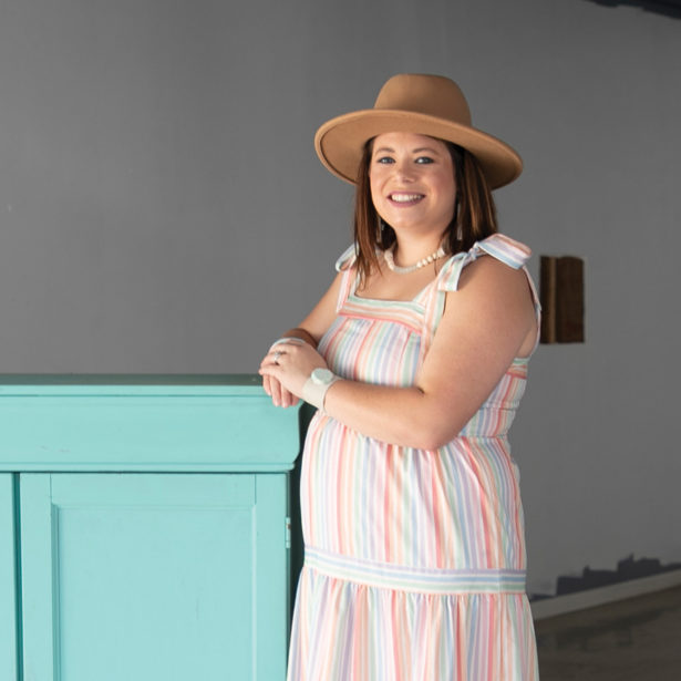 A woman in a seersucker dress and felt hat stands next to a large turquoise chest of drawers