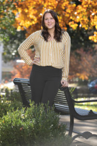 A woman in a striped shirt stands under a autumn tree next to a park bench