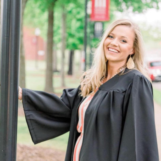 A smiling blonde woman in a black robe stands by a light post on the UT Southern campus