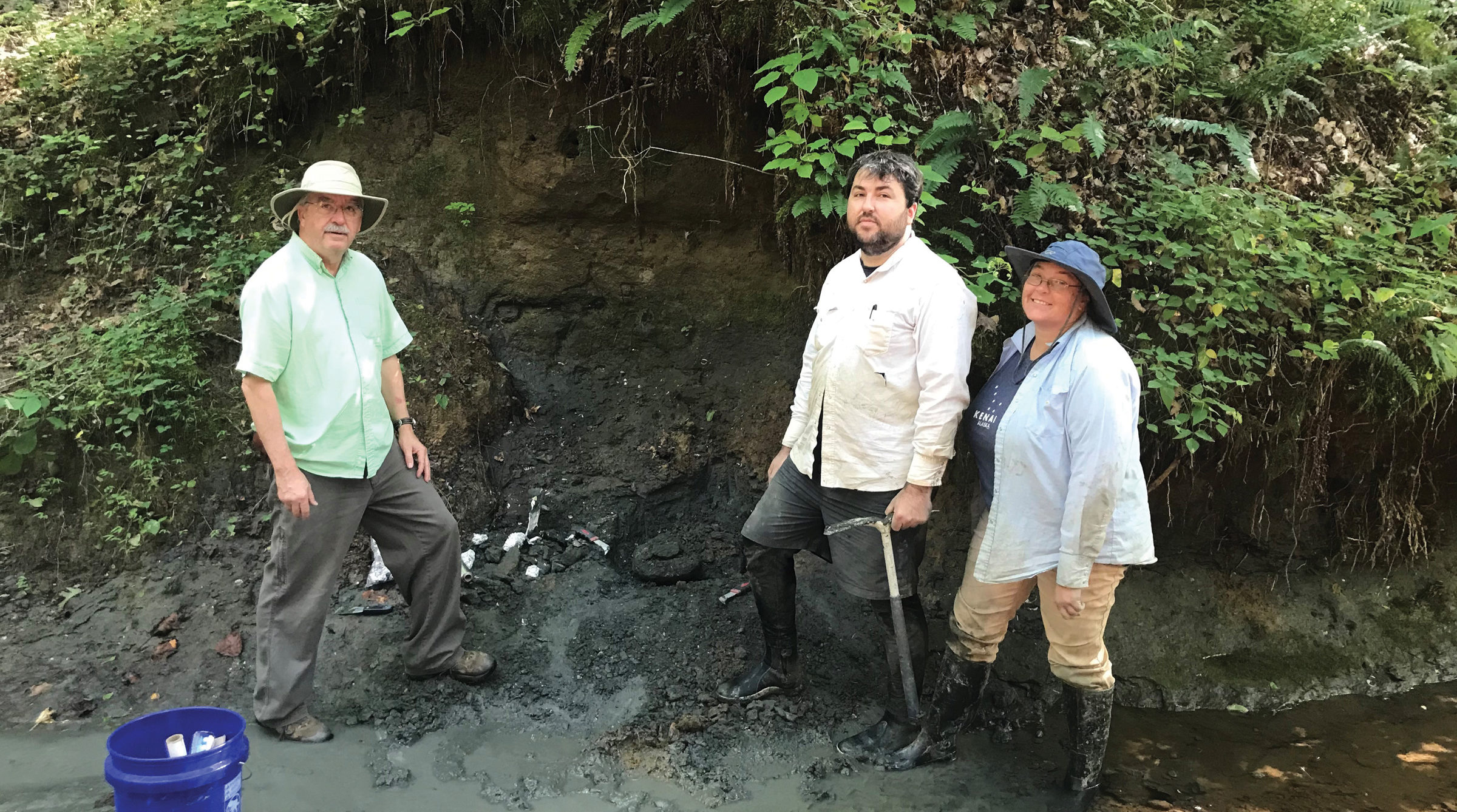 Three researchers stand in a creek bed looking for fossils