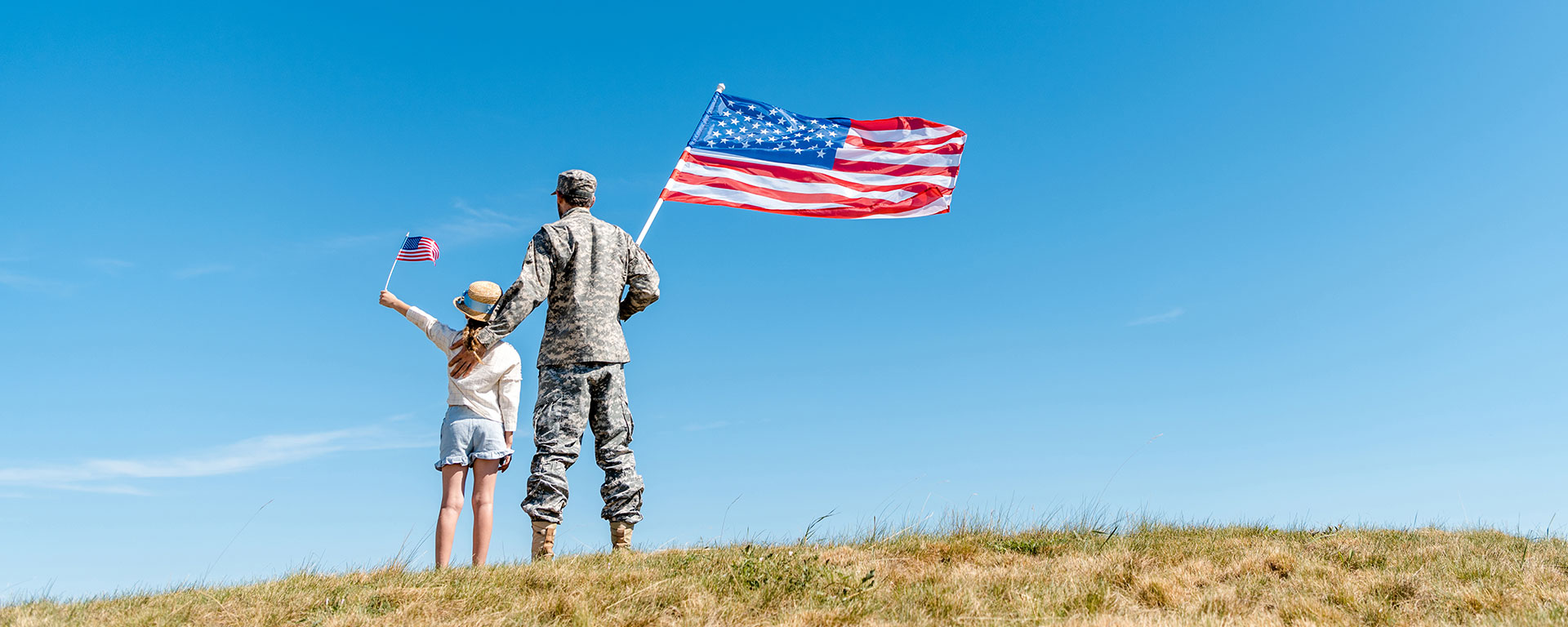 Uniformed military member stands with daughter on a grassy hill, with US flags waving in the wind.