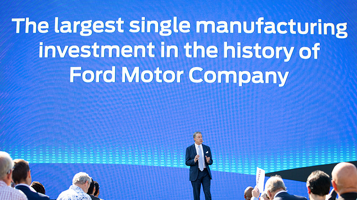 A Ford Motor company employee speaking, with a blue projected screen behind him with the words "The largest single manufacturing investment in the history of Ford Motor Company."