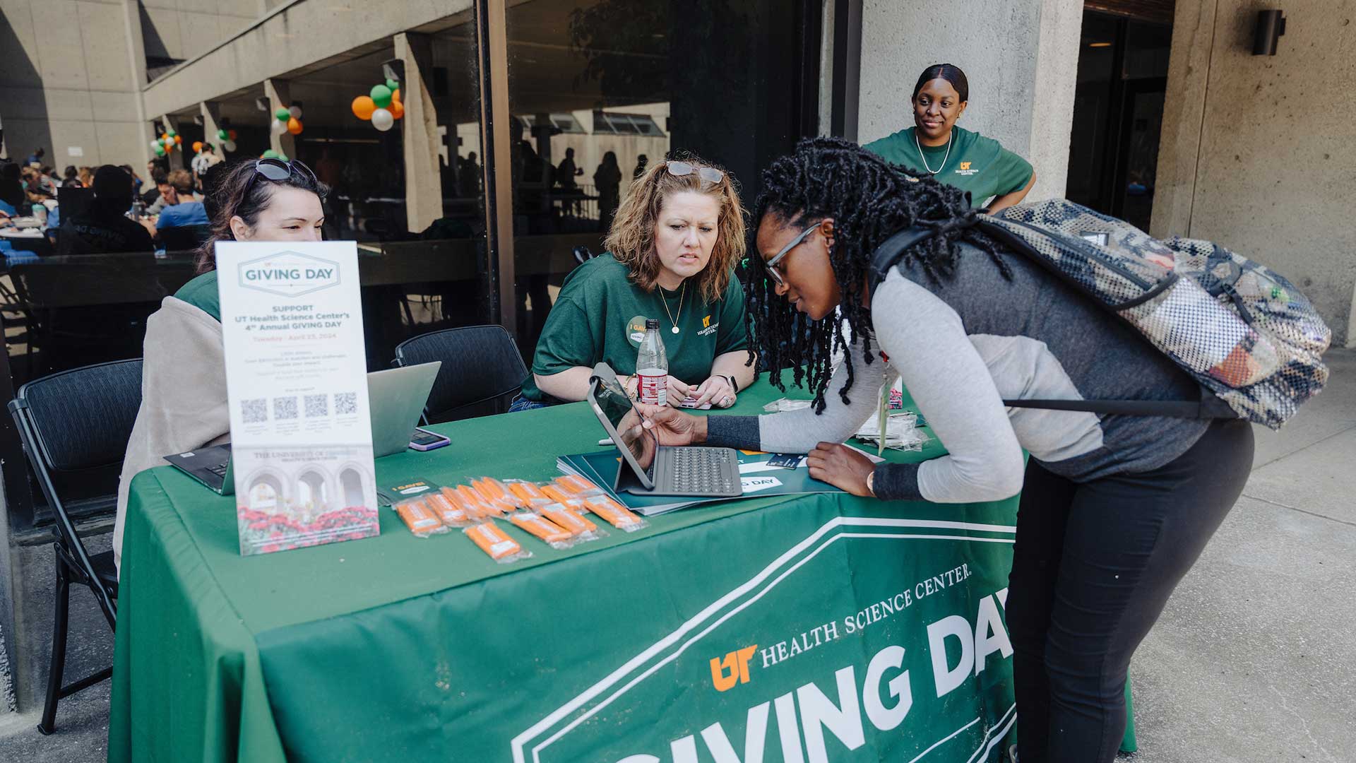 A student using an iPad to make a donation at a UTHSC Giving Day table run by three female workers.