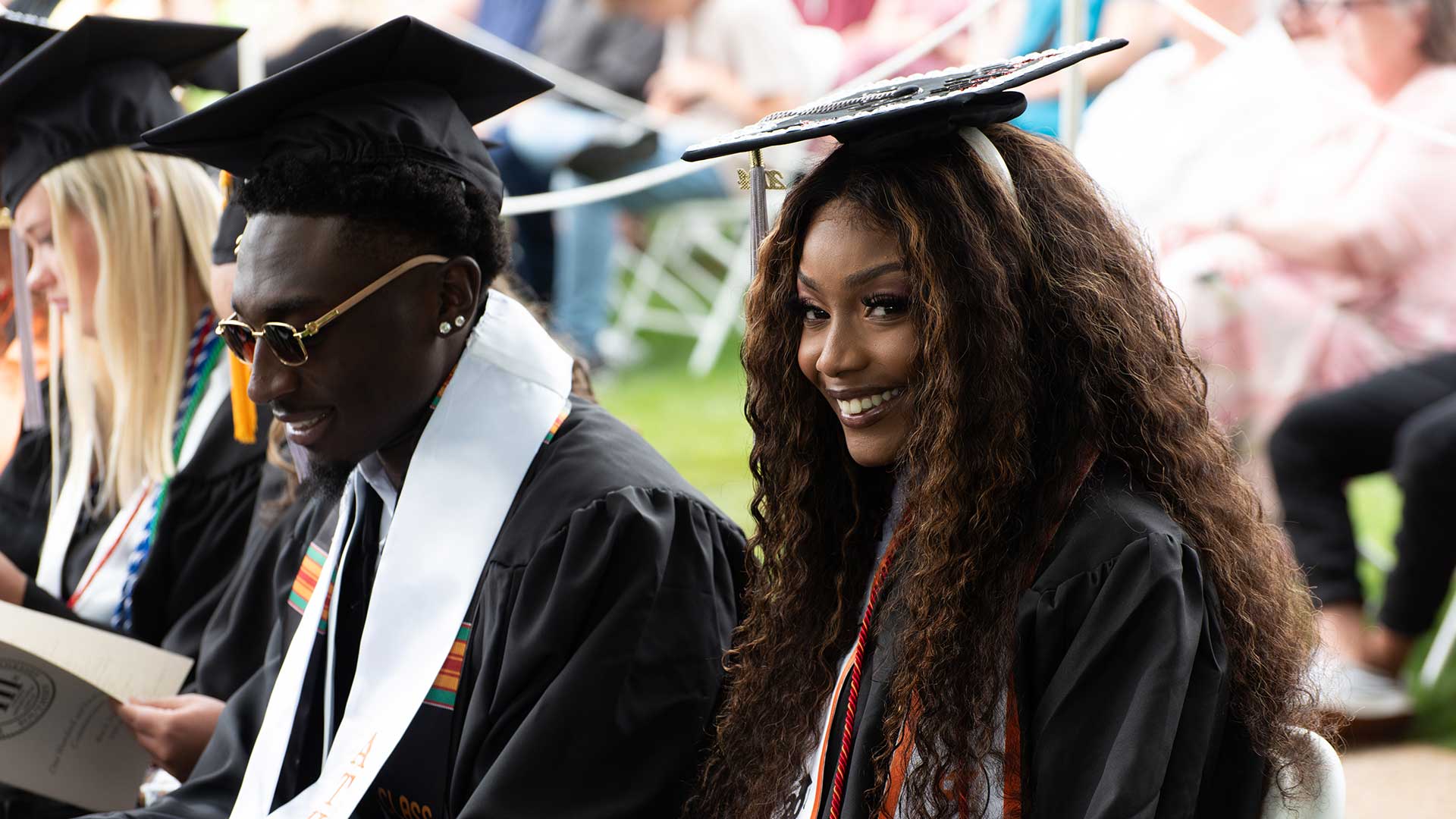 A black female student wearing graduation robes and cap smiles while waiting for the commencement ceremony to begin.