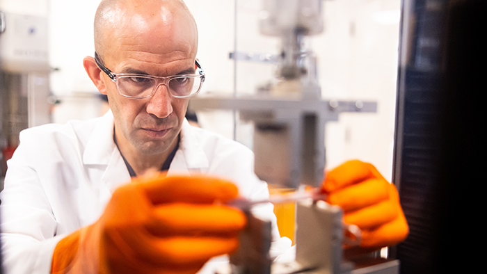 A bald man wearing orange gloves and safety glasses performs scientific testing on a material.
