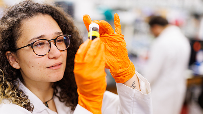 A female scientist wearing orange gloves examines a small contained with a dark substance inside.