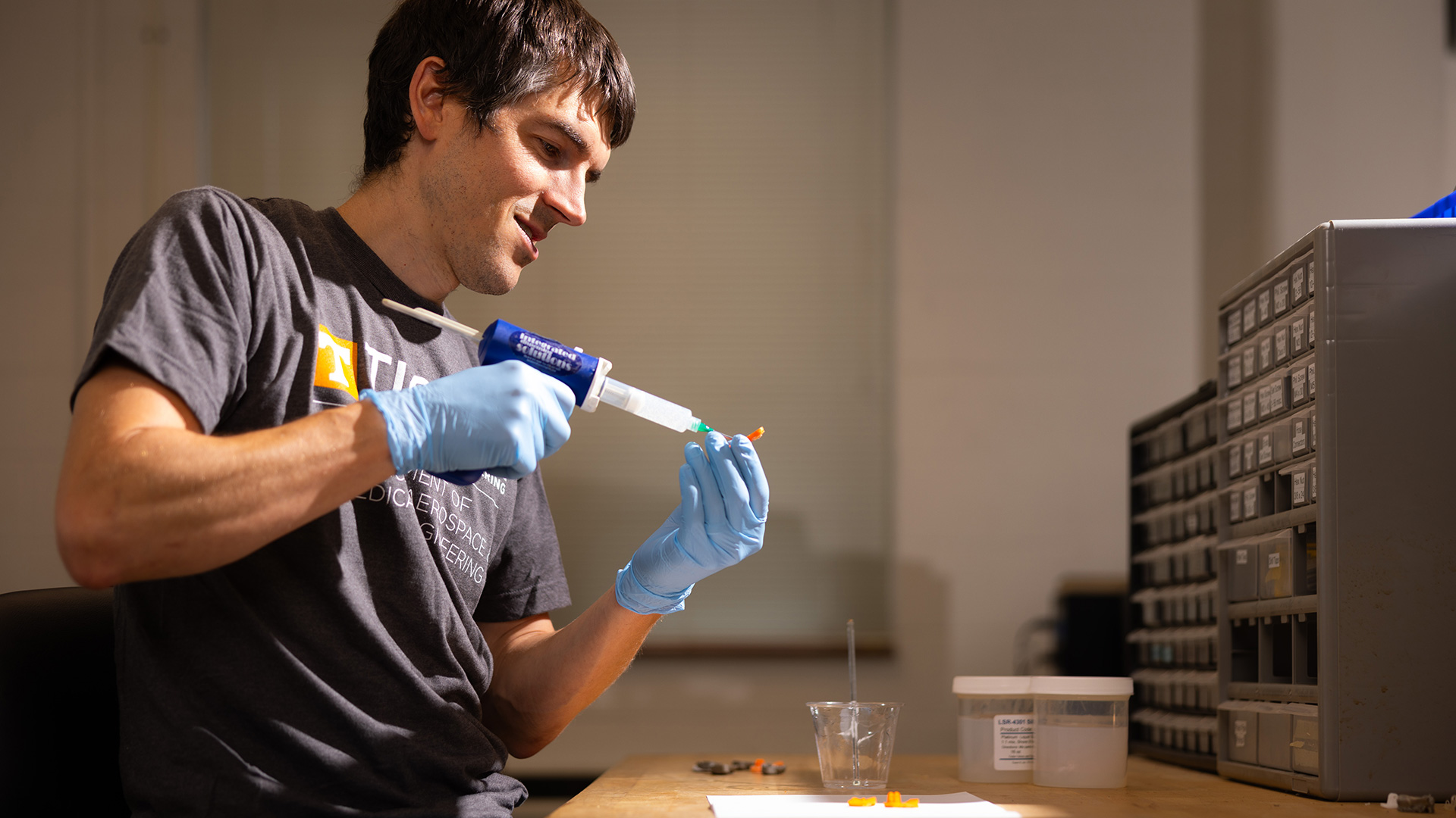 A male student wearing blue gloves applies silicone to a 3D printed part of a prosthetic foot.