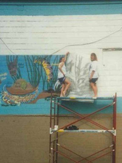 Faded photo of two women standing on scaffolding, painting a mural on the side of a building.